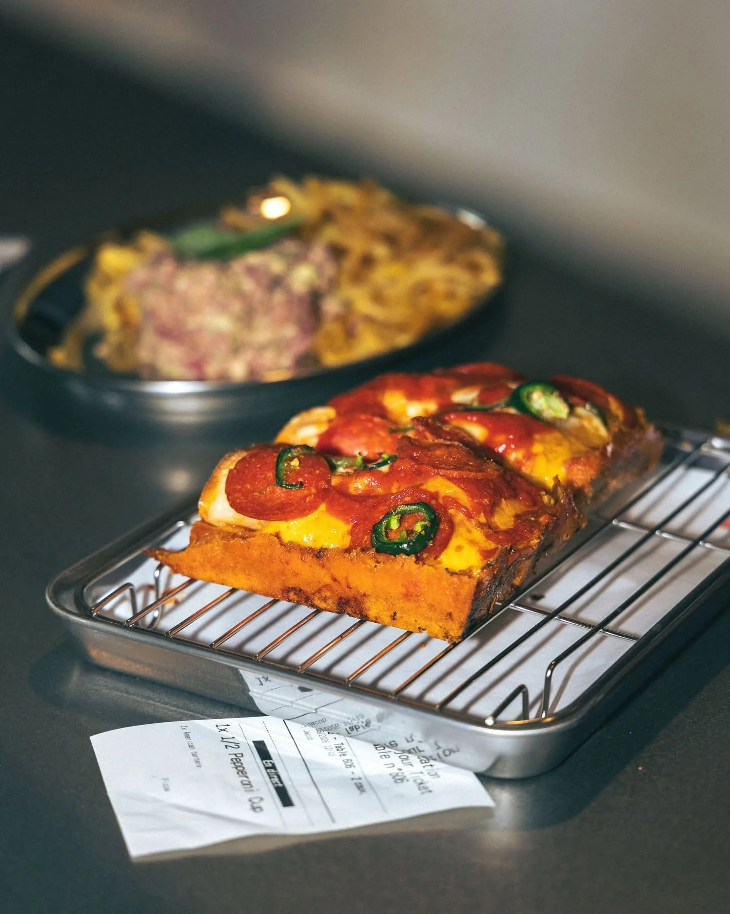 Pizza Detroit pepperoni et jalapeños servie sur une grille, avec un plat de pâtes en arrière-plan, chez Toto's à Rouen.